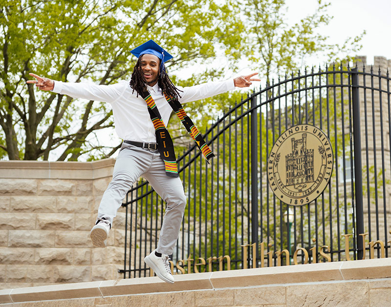Student in a graduation cap jumping in front of the gates to Eastern Illinois University