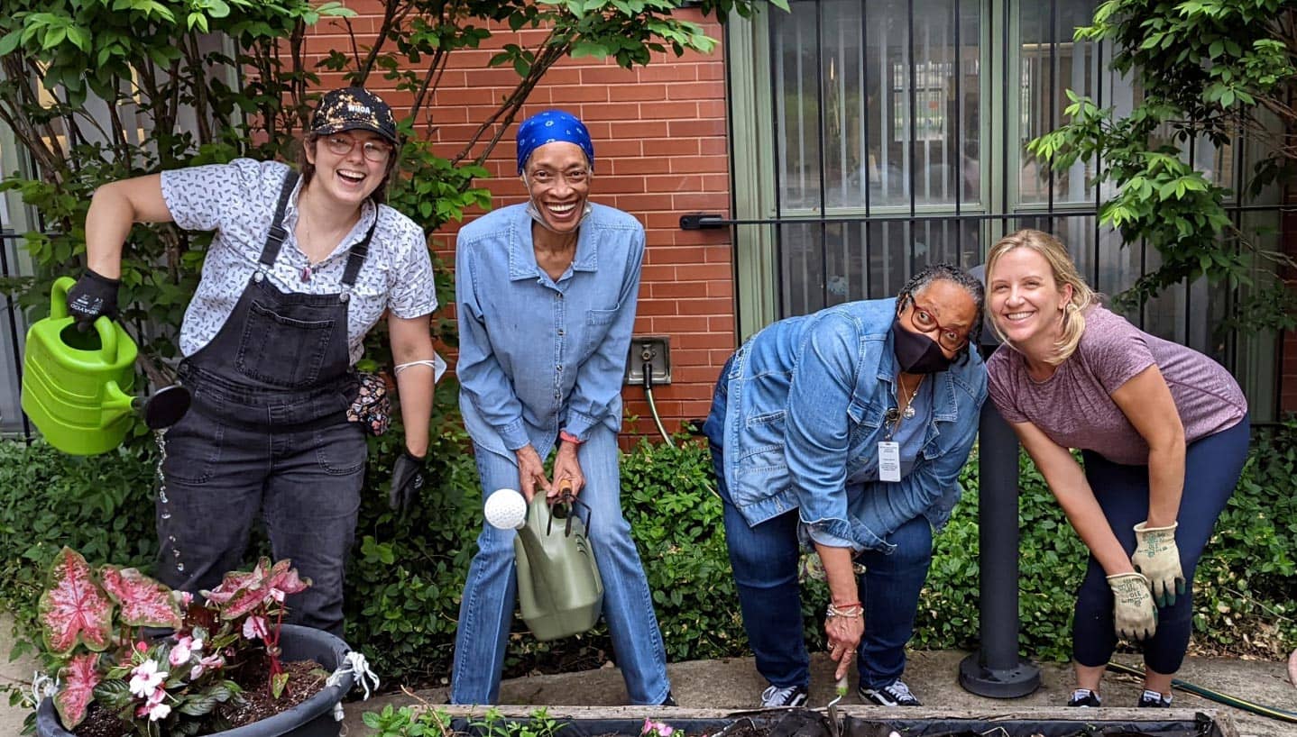 Four women in a community garden holding gardening tools and smiling at the camera.